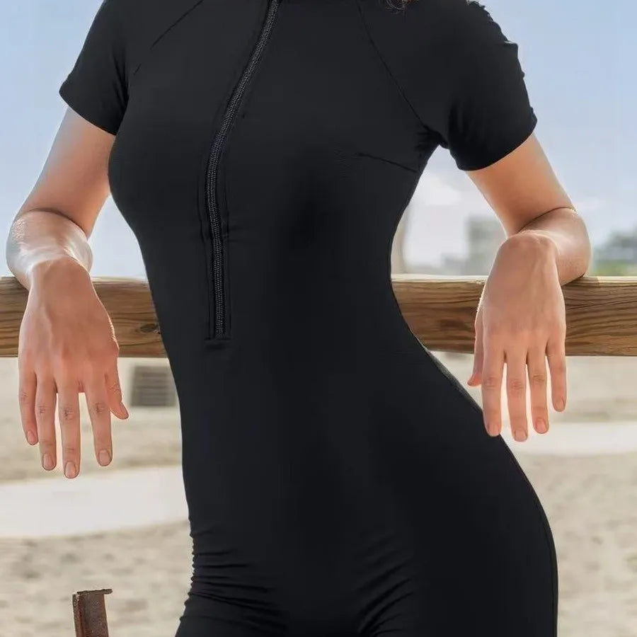 Woman wearing a black short-sleeve one-piece swimsuit with a front zipper at the beach.