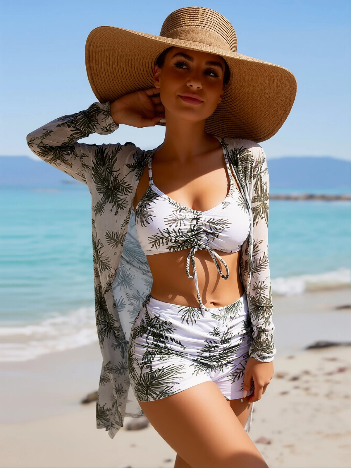 Woman wearing a palm print bikini set and matching cover-up with a wide-brim straw hat on a beach.