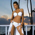 Woman in white bikini set and matching beach cover-up posing by the ocean at sunset.